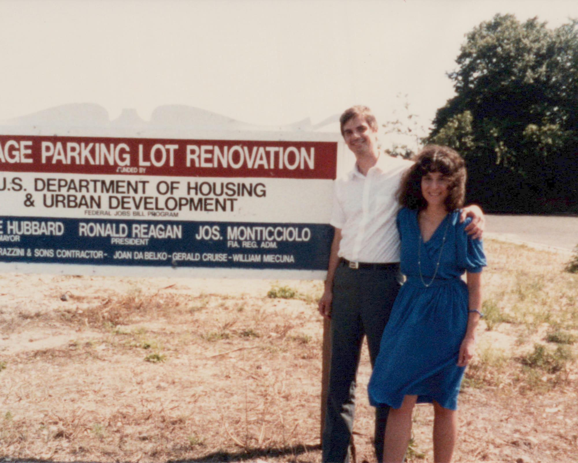 William Miecuna standing with Adele in front of a HUD project sign for a parking lot renovation, his name printed among the officials listed