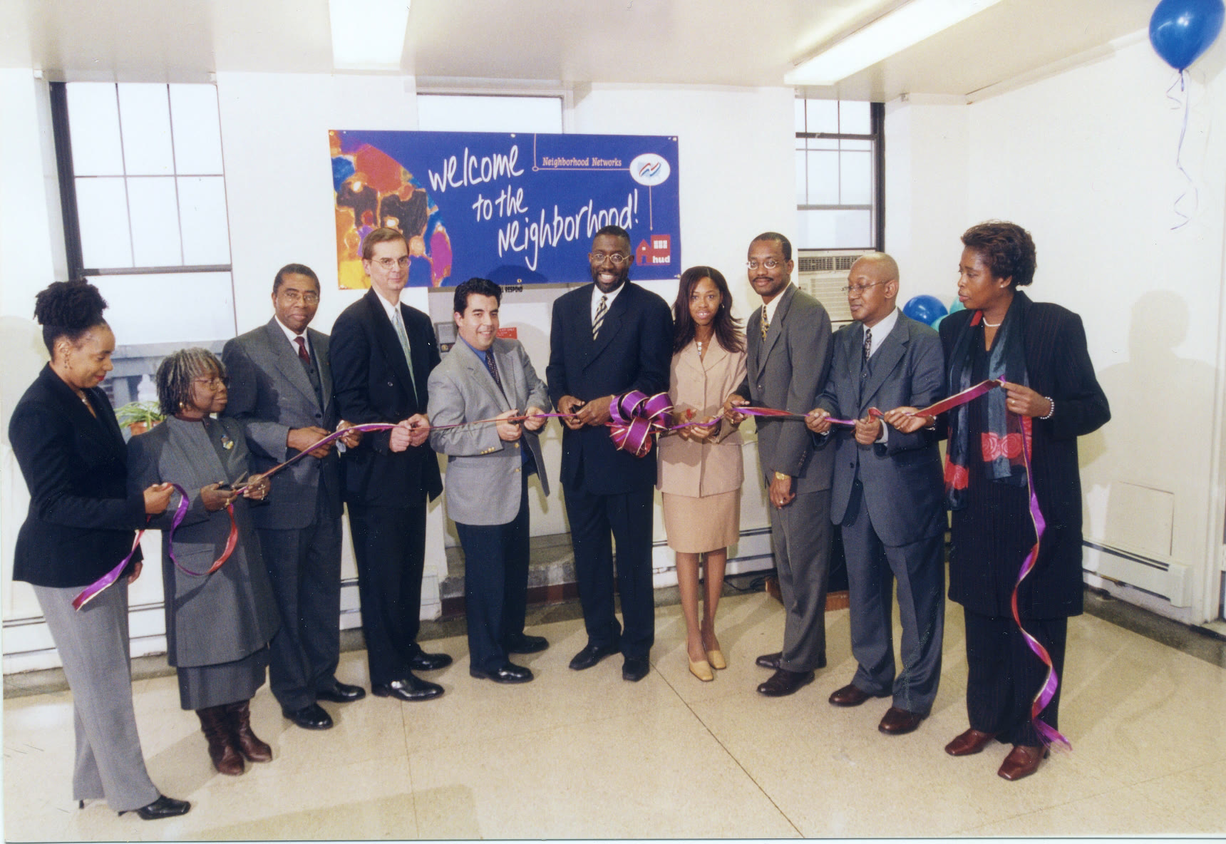 A long line of officials and residents cutting a ribbon beneath a HUD 'Welcome to the Neighborhood' banner